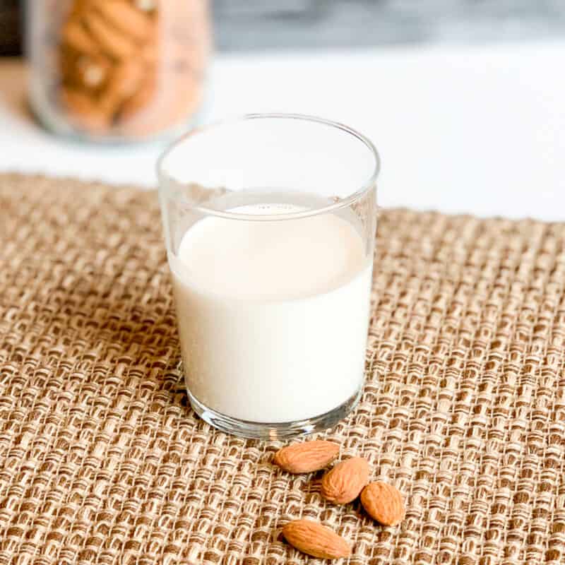 side view of homemade almond milk in a glass sitting on a woven brown placemat next to lose almonds