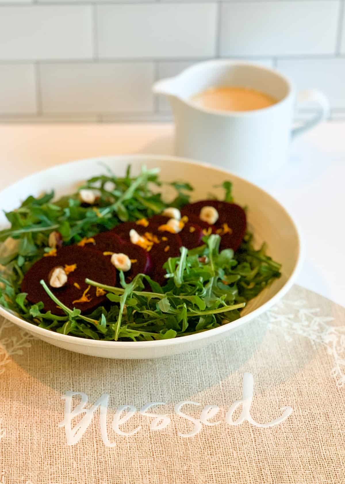 Elegant Beet Arugula Salad in a white bowl sitting on a placemat that reads "Blessed" and a blurred container of orange vinaigrette dressing in the background
