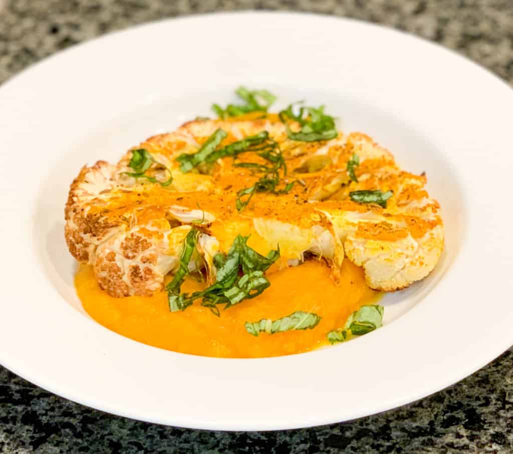 close up of prepared cauliflower steak with butternut squash puree topped with fresh basil in a white wide rimmed bowl