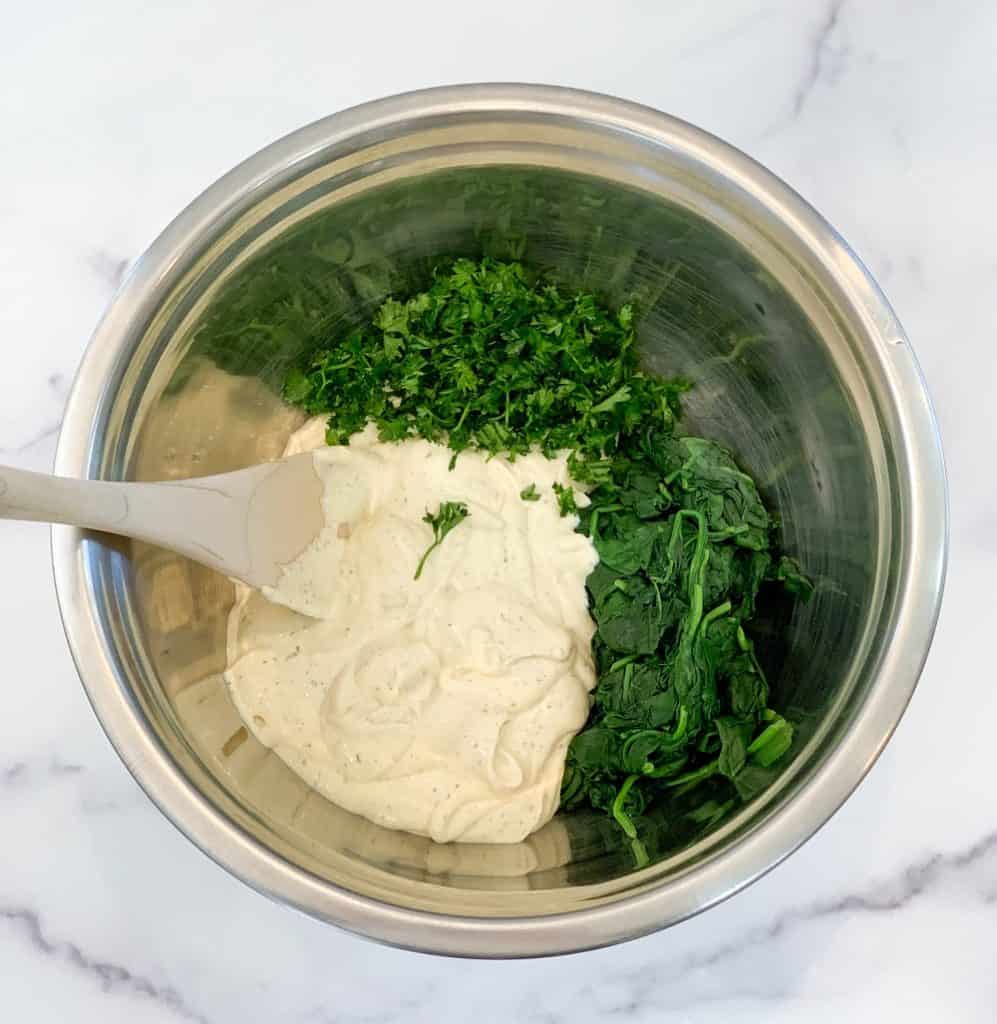 Top view close up of a stainless steel mixing bowl with tofu ricotta, sautéed spinach and fresh parsley sitting a marble looking surface