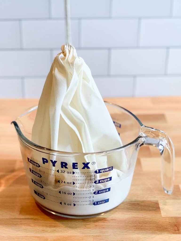 The nut milk bag with almond milk being lifted from the large measuring cup.