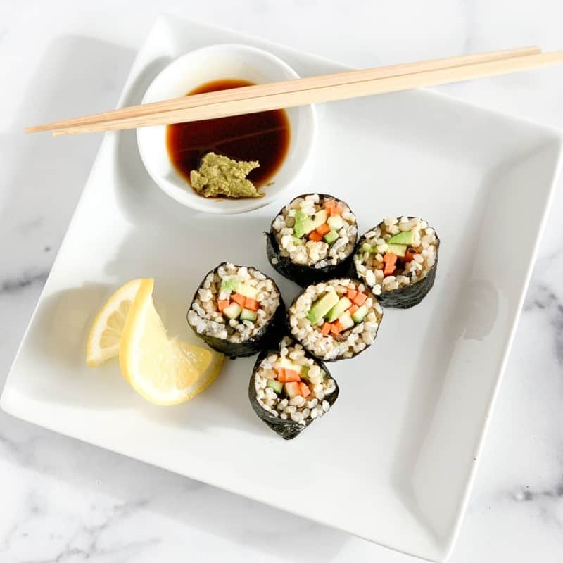 top view close up of veggie sushi rolls on a square white plate with slice of lemon and dish with soy sauce and wasabi