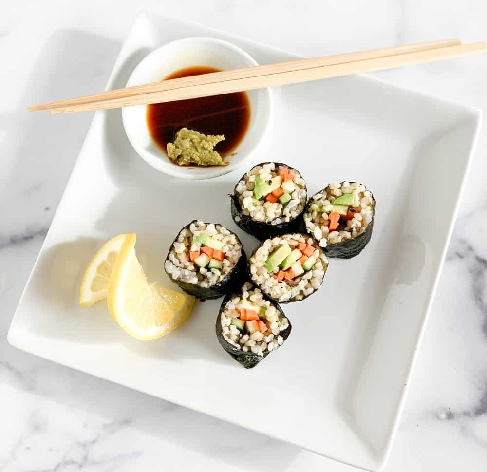 Top close up view of five avocado veggie rolls on a square white plate, a side of soy sauce and wasabi paste, lemon slice, and a pair of wooden chopsticks.