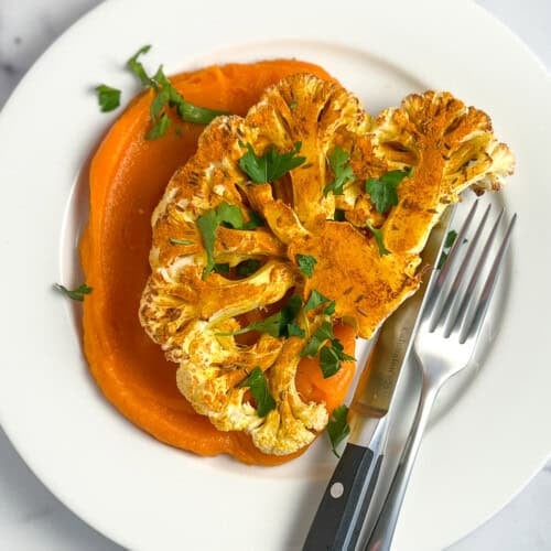 top view of a spiced cauliflower steak on top of butternut squash puree with fresh chopped parsley sprinkled on top; steak knife and fork on the side