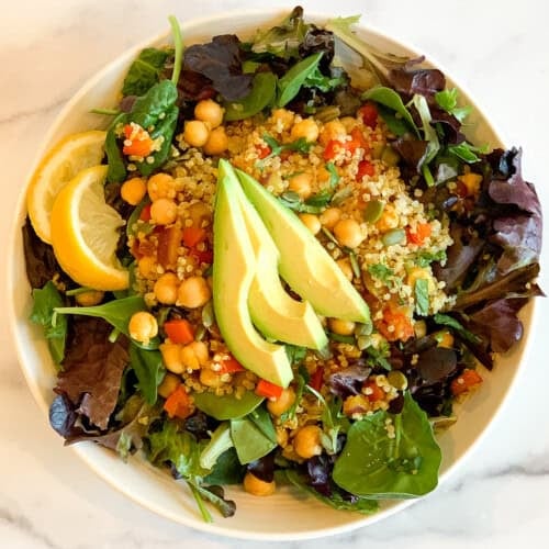 A bowl of curry quinoa chickpea salad with sliced avocado and two lemon wedges.