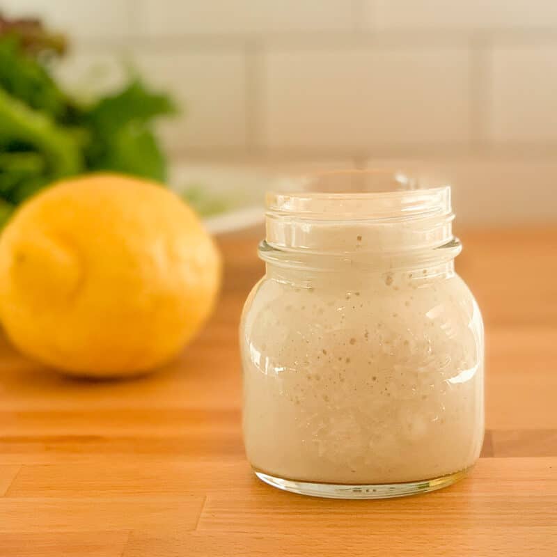 Side view close up of tahini lemon sauce in a glass jar with lemon and salad greens blurred in the background