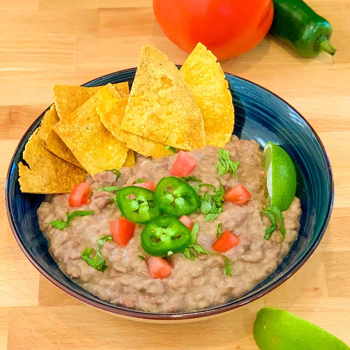 top view of oil free refried beans in a dark blue bowl with oven baked tortilla chips, and chopped tomatoes, cilantro and jalapeno. lime wedges on the side