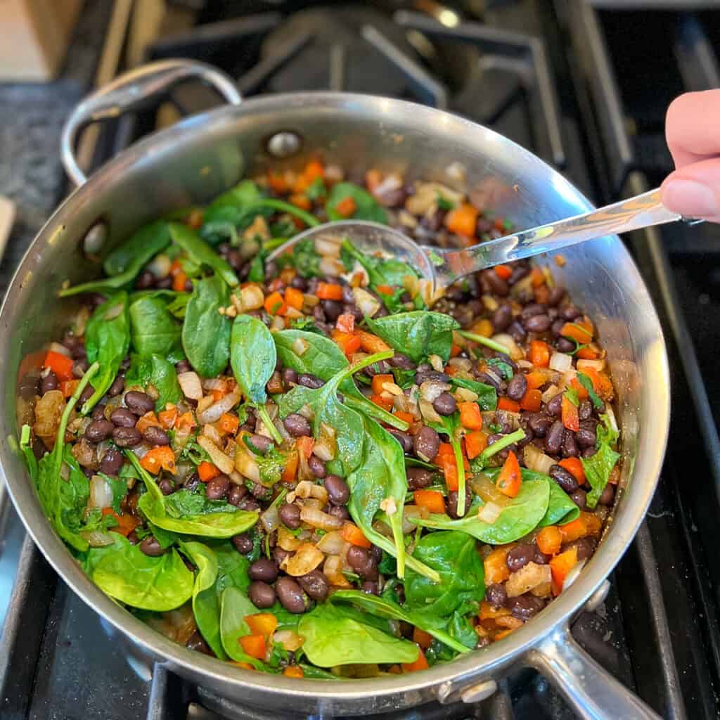 A woman's hand using a spoon to stir the spinach into the bean and veggies mixture in the sauté pan.