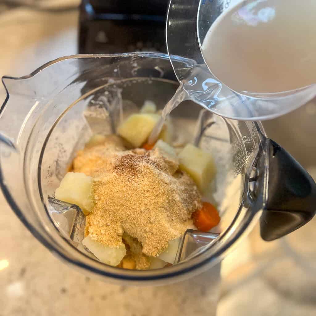 Vegan cheese sauce ingredients being added to a blender pitcher with reserved cooking water being poured in.
