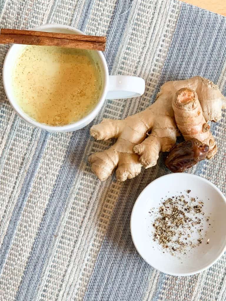 the top view of the golden milk latte in a white mug alongside the key ingredients of turmeric, ginger, date, black pepper in the latte. the latte and ingredients are on top of a striped blue placemat.