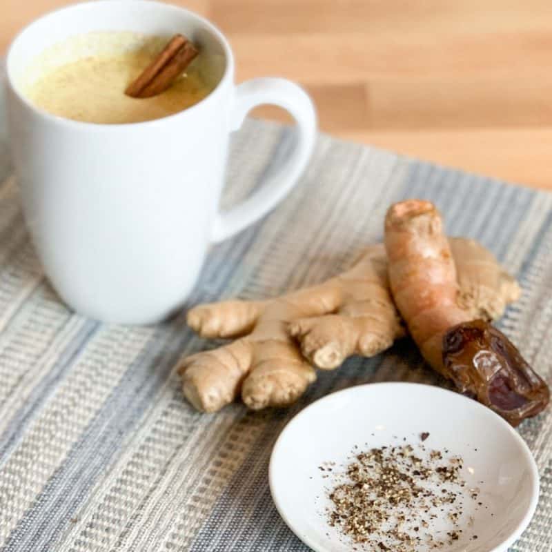 golden milk latte in a white mug alongside the key ingredients of turmeric, ginger, date, black pepper in the latte. the latte and ingredients are on top of a striped blue placemat.