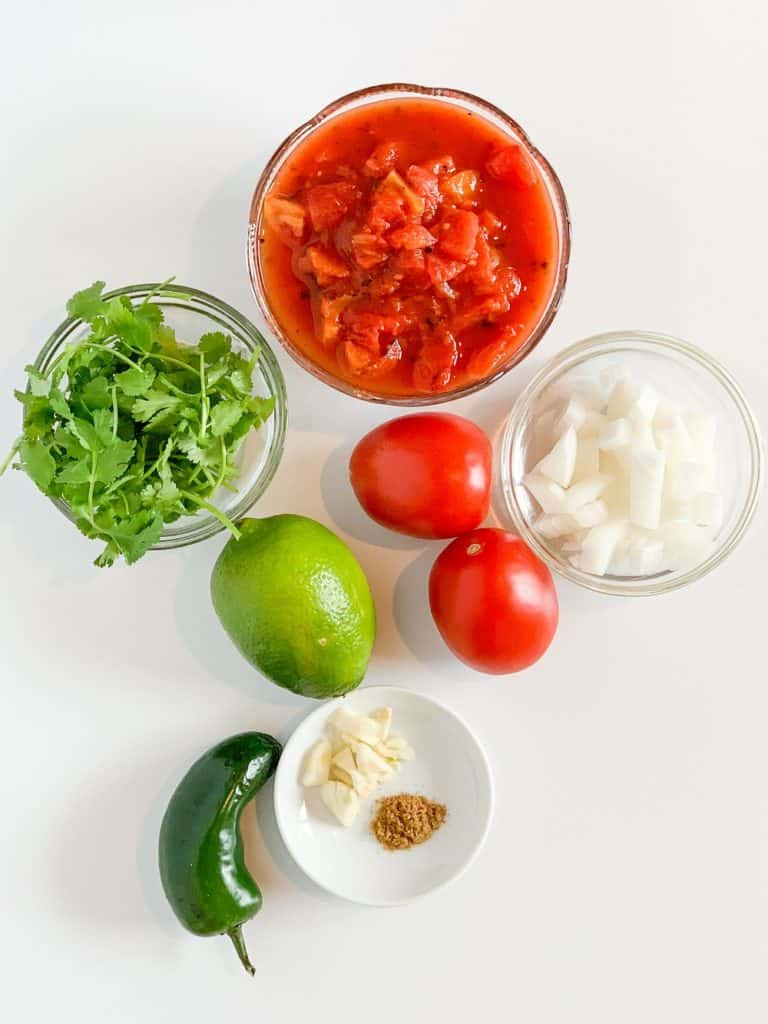top view of ingredients for salsa: fire roasted tomatoes, cilantro, onion, Roma tomatoes, lime, garlic, cumin, jalapeno
