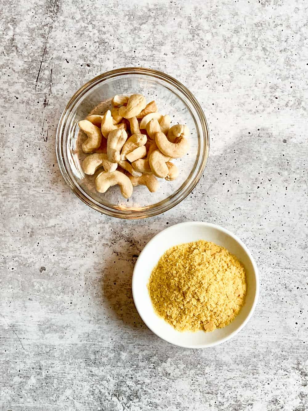 The top view and close up of a small glass bowl of cashews next to a smaller white bowl of nutritional yeast--against a rough grey backdrop.