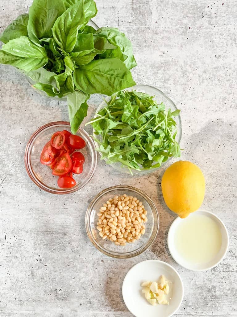 a top view of the ingredients for arugula basil pesto sauce against a grey background. fresh basil, arugula, cherry tomatoes, pine nuts, lemon juice and chopped garlic
