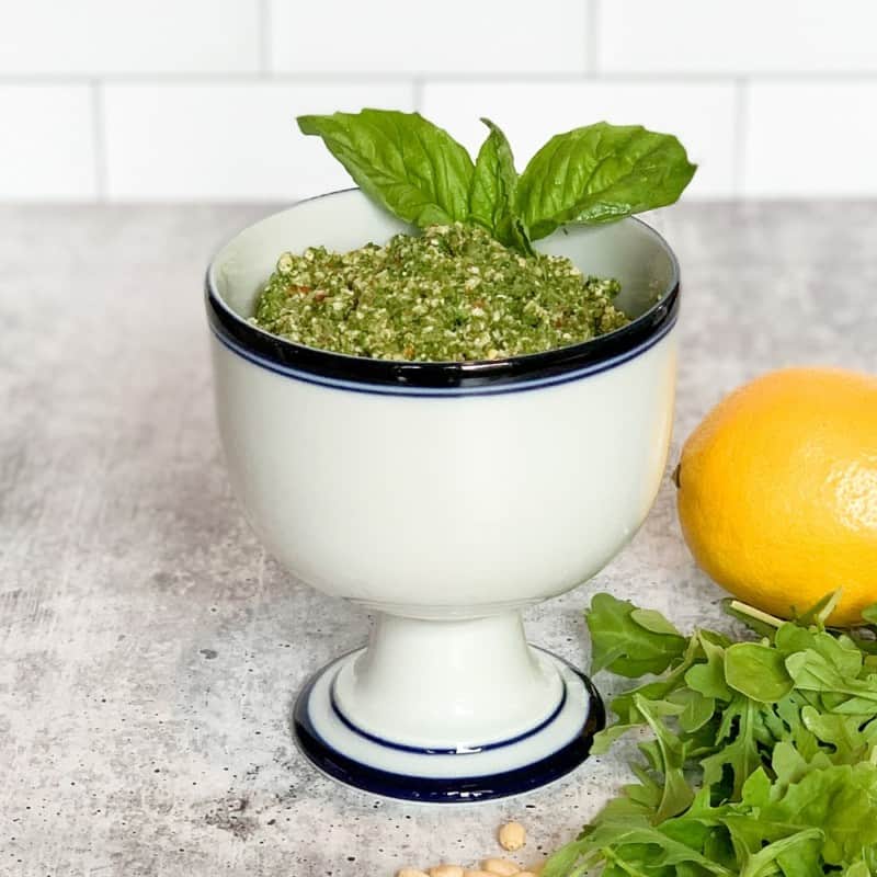 a white pedestal bowl with dark blue rim filled with arugula basil pesto sauce, next to a lemon, arugula and pine nuts on a grey surface and white tiled backgroud