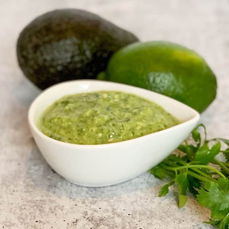 a small white ceramic bowl of avocado cilantro lime dressing next to an avocado, lime and springs of fresh cilantro