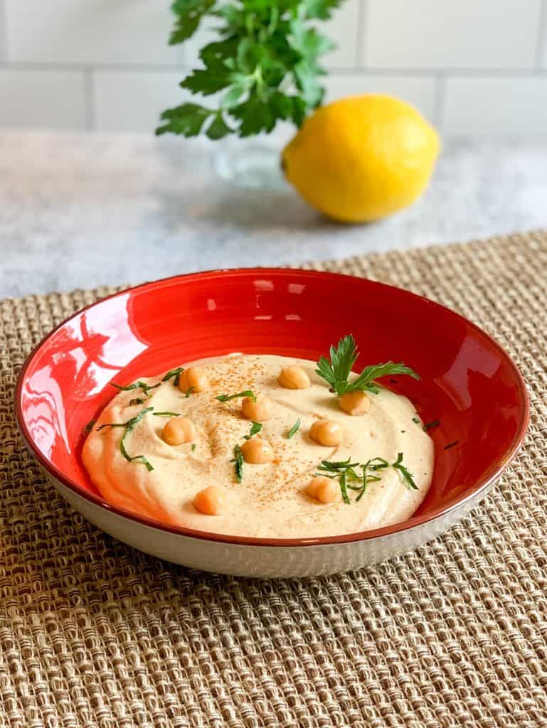 Best Garlic Hummus in a red bowl on a woven mat with fresh parsley on top and blurred in the background next to a lemon