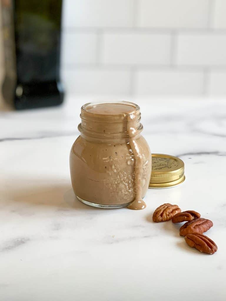 Clear jar with the best toasted pecan balsamic vinaigrette on marble surface with four pecan halves in front
