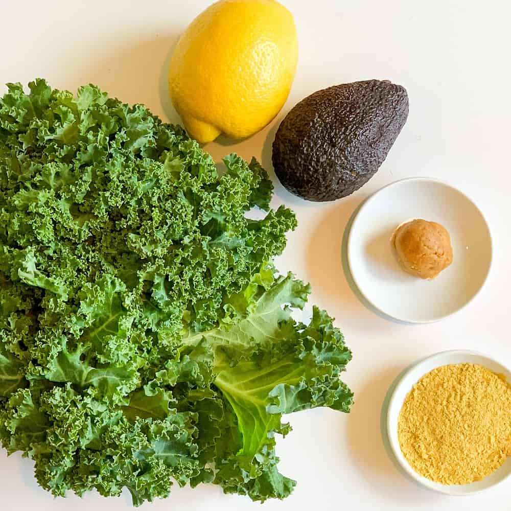 ingredients for yummy avocado and kale salad - fresh curly kale, lemon, miso paste, nutritional yeast and avocado against a white background