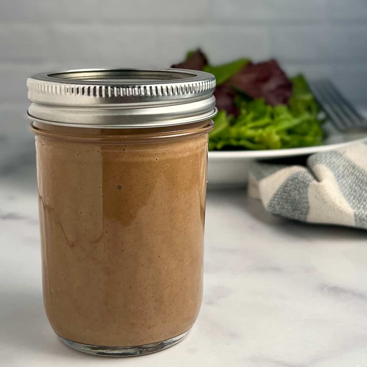 side view of balsamic salad dressing in a mason jar; bowl of salad greens blurred in background