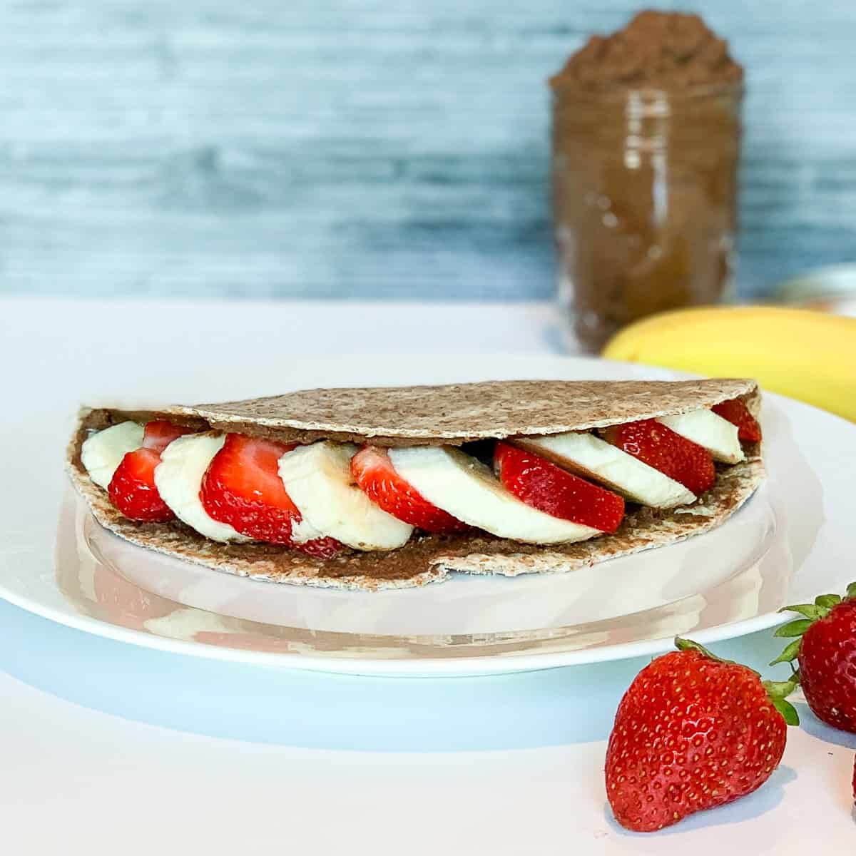 Chocolate hummus breakfast wrap on a plate with strawberries and banana on the side.