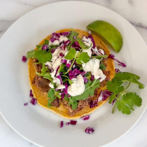 Refried bean tostadas topped with veggies and cashew cream on a plate with lime wedge.