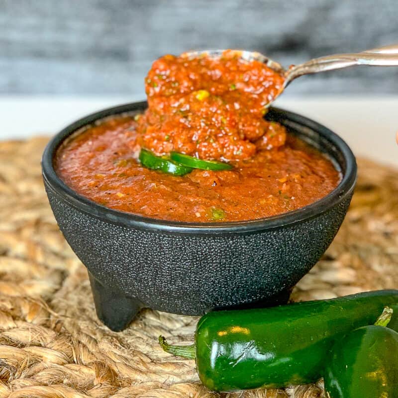 side view close up of red salsa in a bowl with fresh jalapeno and a spoon dishing some out