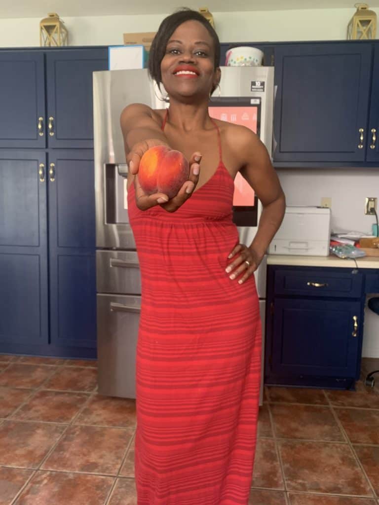Full view of Sersie in a red dress in a kitchen holding a fresh peach out front.