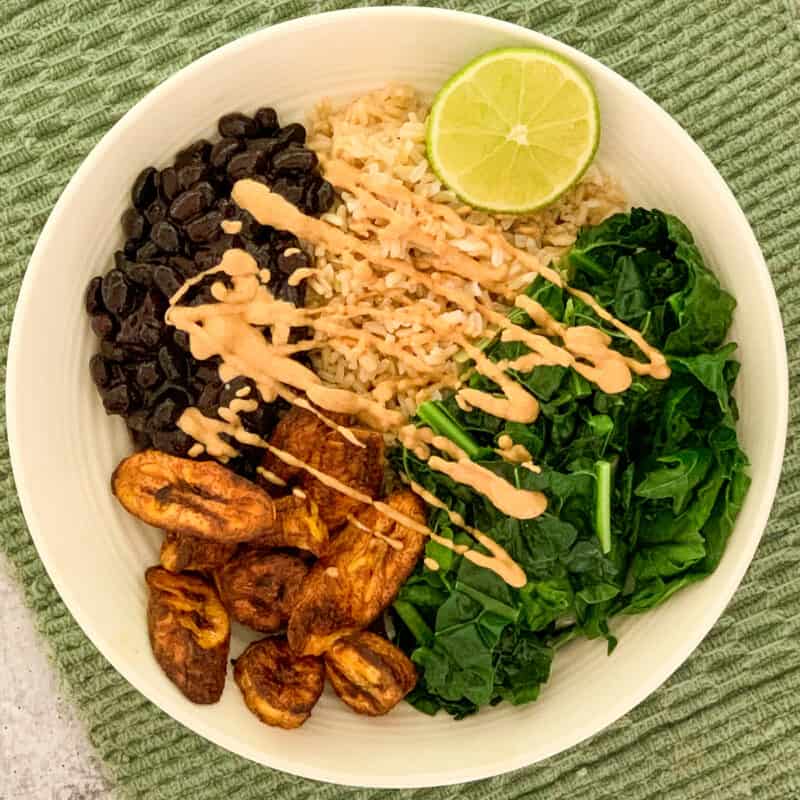 top view close up of black bean buddha bowl with black beans, brown rice, steamed kale, air fryer plantains, lime wedge and chipotle sauce drizzled on top