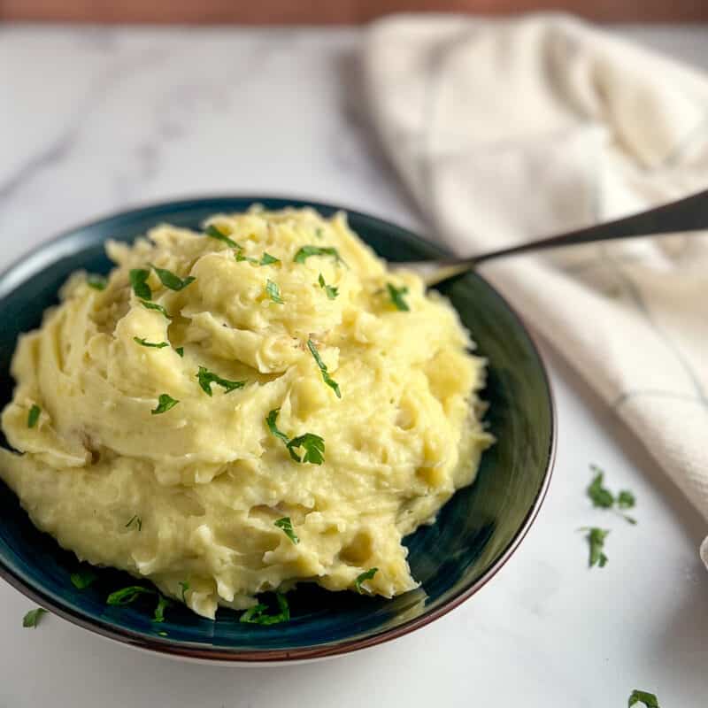 side view of garlic mashed potatoes in a blue bowl with spoon sticking out. napkin blurred in the background.
