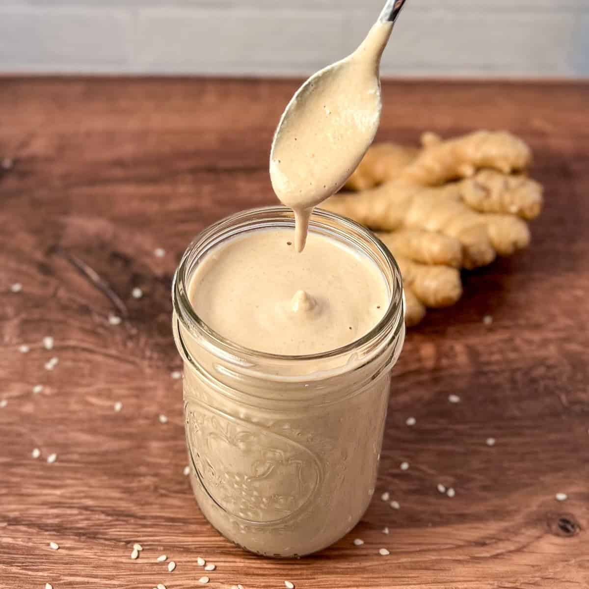 Creamy tahini ginger dressing in a mason jar with a spoon lifting some out; ginger root blurred in the background with scattered sesame seeds.
