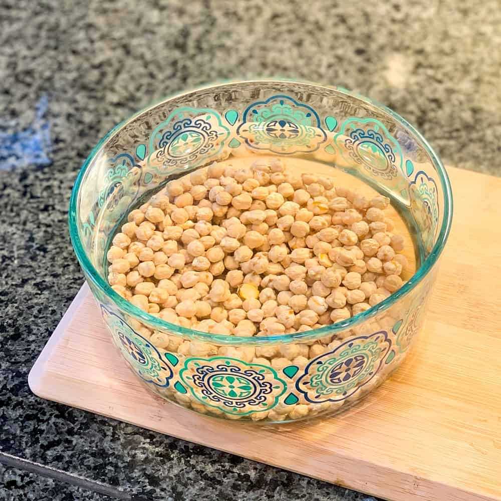 decorative glass bowl container with dried chickpeas soaking in water sitting on a wooden cutting board for The Best Falafel That Happens to be OIL-FREE.