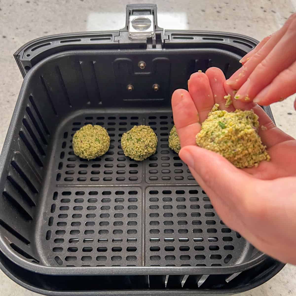 A woman's hand forming falafel patties and placing them in the air fryer basket.