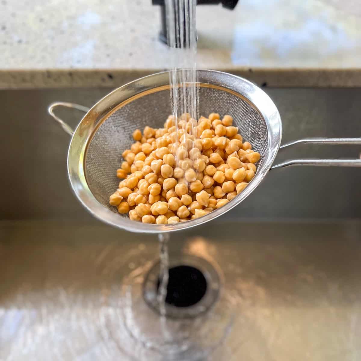 Soaked chickpeas being drained and rinsed in a kitchen sink with a colander.