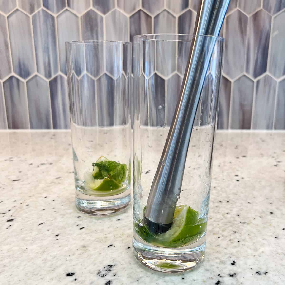 Two tall glasses with fresh mint and lime, one glass has a muddle inside.