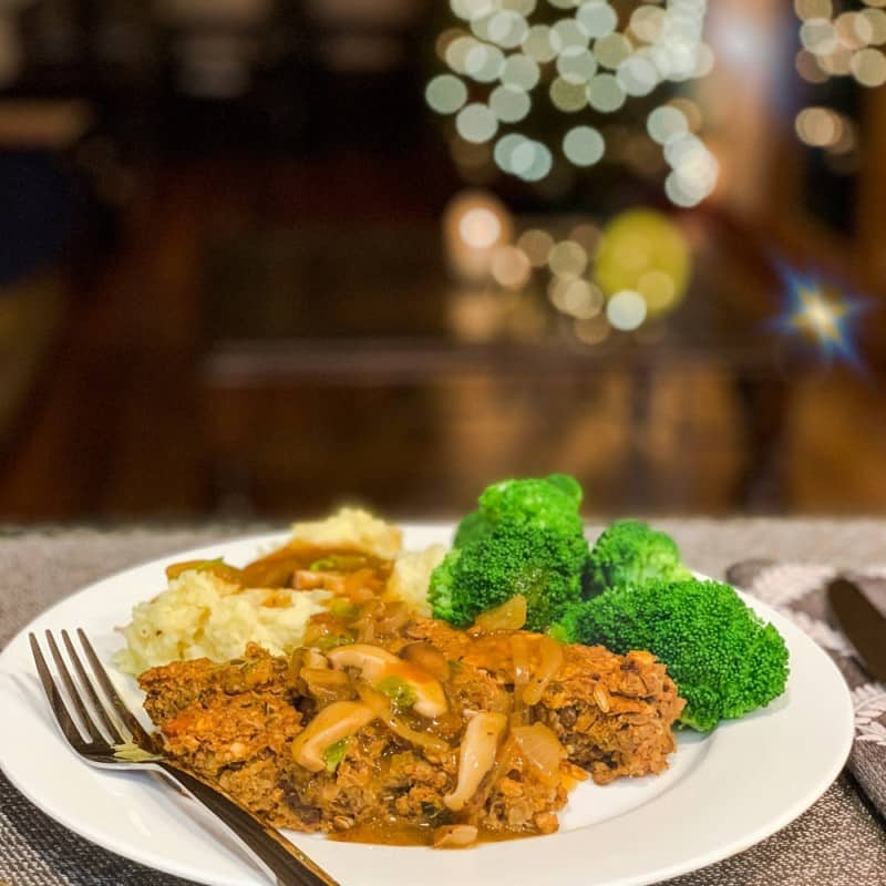 side view of vegan lentil loaf with mashed potatoes, mushroom gravy, steamed broccoli and a Christmas tree blurred in the background
