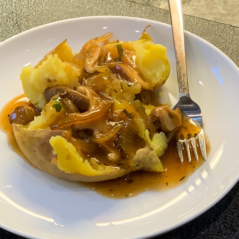 Close of a smashed baked potato with a generous helping of caramelized onion and mushroom gravy on a white plate with a small fork next to it. 