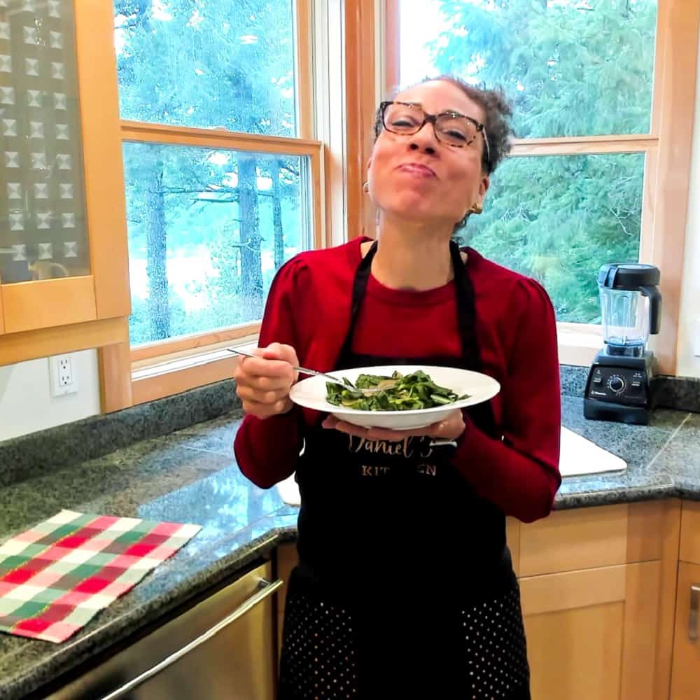 Gigi standing in her kitchen, smiling and eating a bowl of Southern Style Collard Greens (vegan)