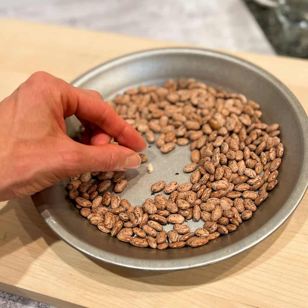 a hand sorting through dried pinto beans in a pie tin sitting on a wooden cutting board