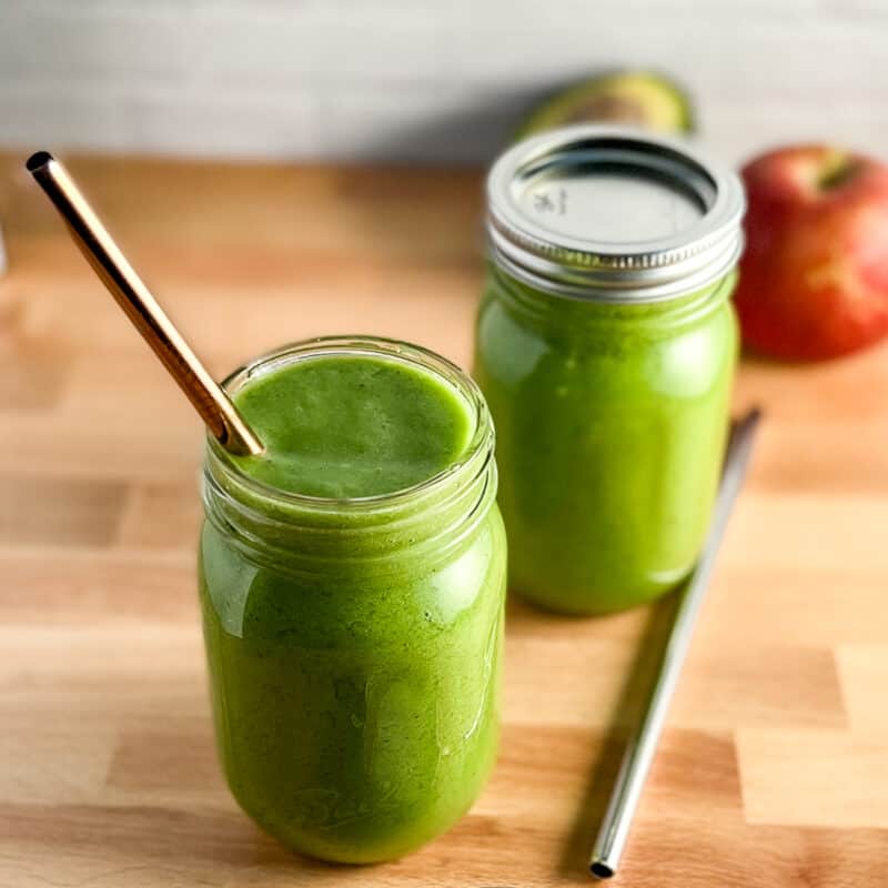 side and top view of simple 5-ingredient smoothie in two glass mason jars with reusable metal straws sitting on a wood surface. apple and avocado are blurred in the background