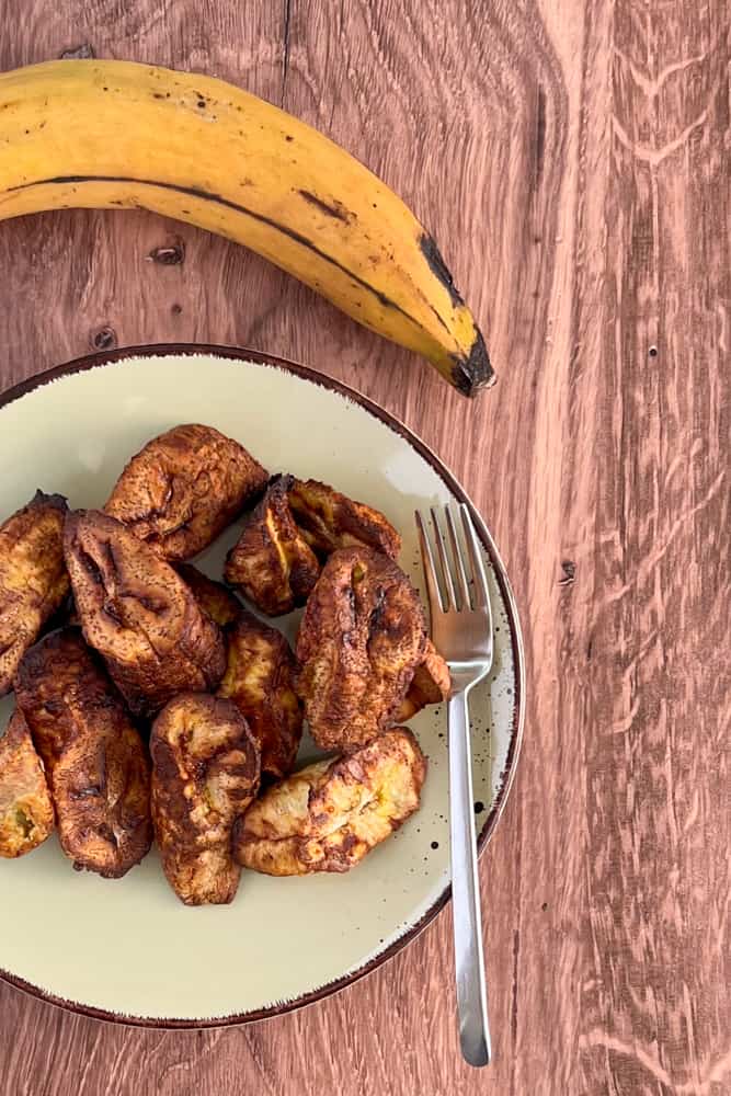 top view of a plate of oil free plantains cooked in an air fryer sitting on a light green plate against a wood surface. A perfectly ripe plantain positioned above the plate of cooked plantain.