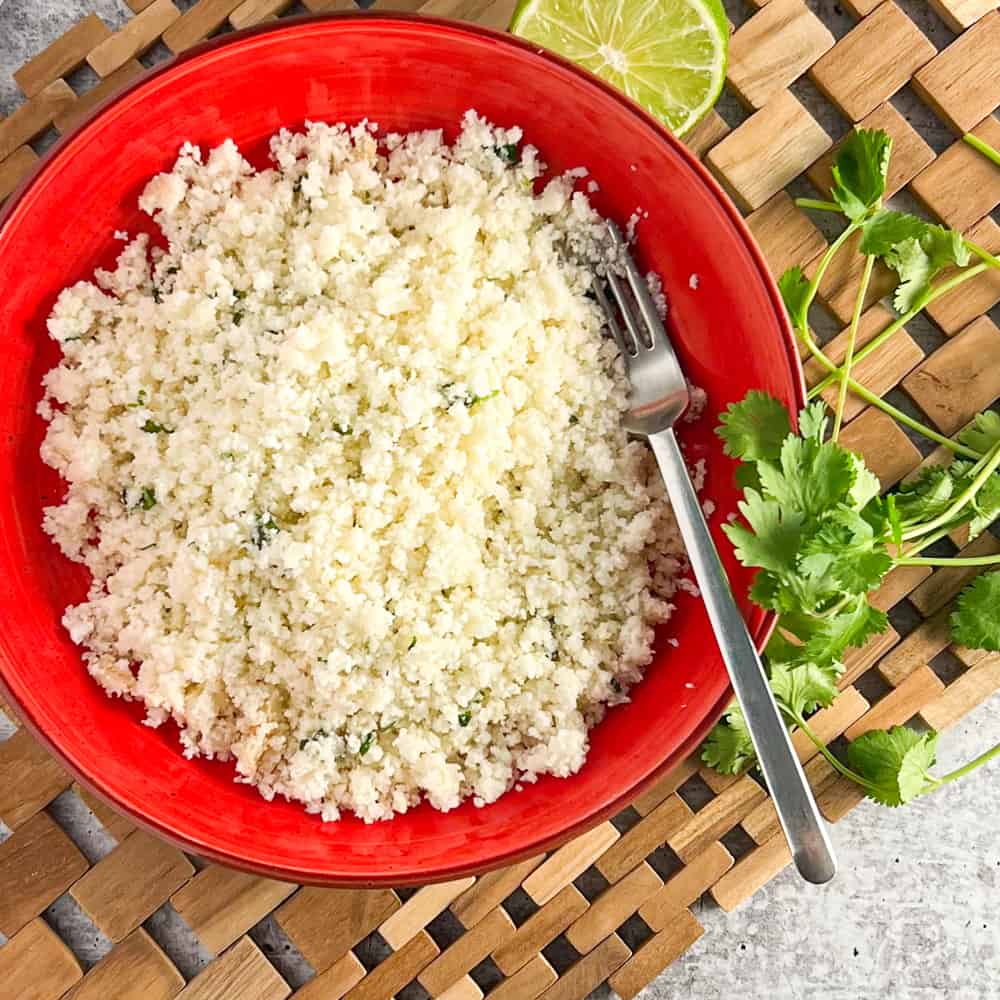 top view of cilantro lime cauliflower rice in a red bowl with fresh cilantro and lime wedge