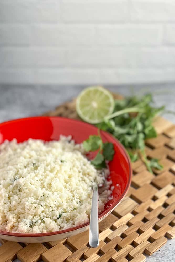 side view of a bowl of cilantro lime cauliflower rice in a red bowl on a wooden place mat with fresh cilantro and lime wedge blurred in the background
