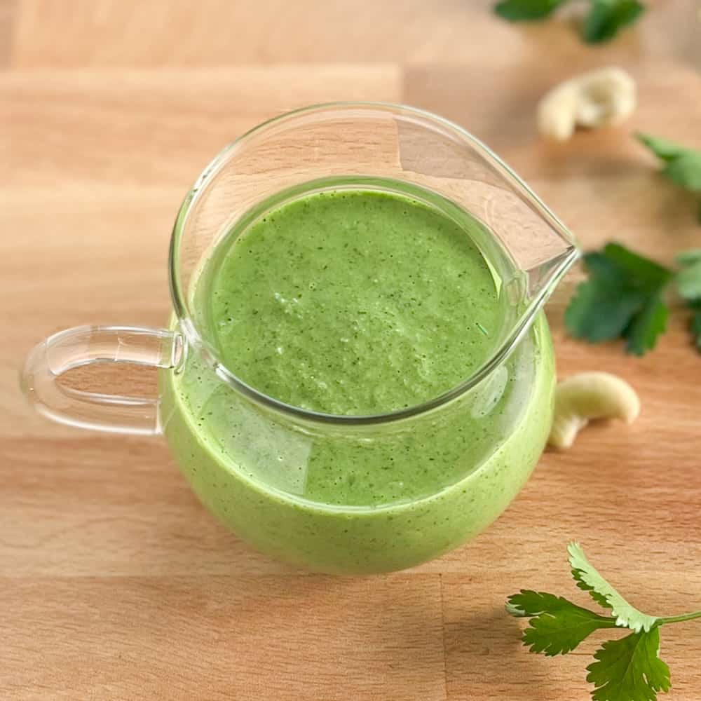 top and side view of easy green goddess dressing in a glass container on a wooden surface with fresh cilantro and cashews blurred in the background