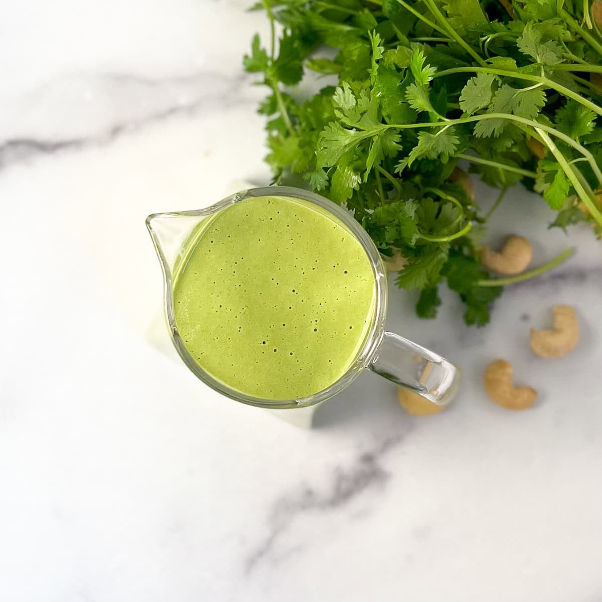 Green goddess dressing in a glass pitcher with fresh herbs and cashews in the background.