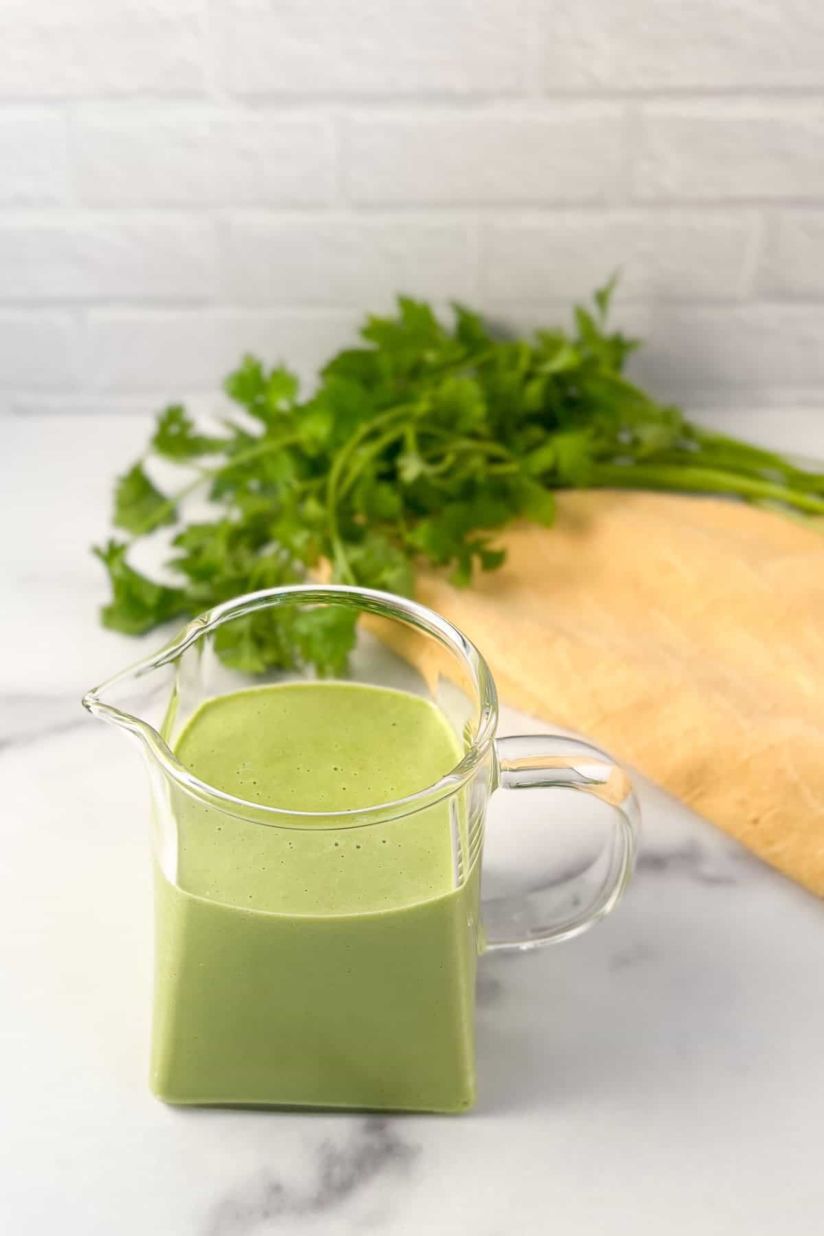 Green goddess dressing in a glass pitcher with fresh herbs in the background.