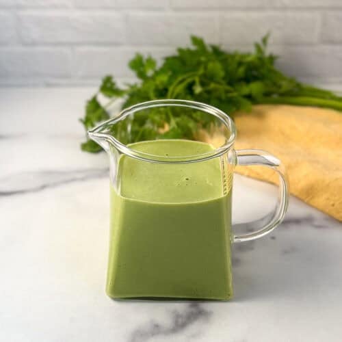 Green goddess dressing in a glass pitcher with fresh herbs in the background.