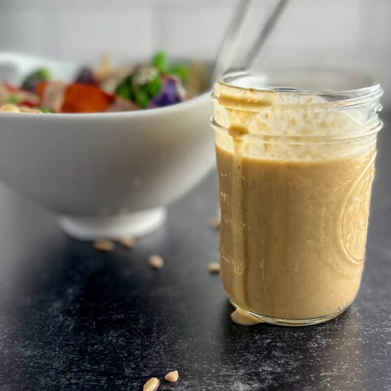side view close up of sweet and tangy sunflower butter sauce in a glass mason jar. a veggie stir fry bowl blurred in the background