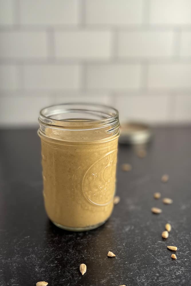 side view of an open mason jar with sweet and tangy sunflower butter sauce on a black surface with lid blurred in the background. loose sunflower seeds scattered on the surface.