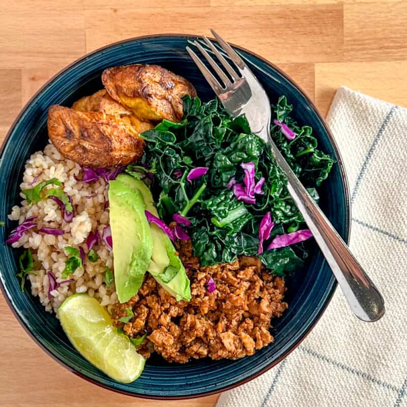 top view close up of sofritas bowl with cilantro brown rice, plantain, steamed kale and sliced avocado in a dark blue bowl sitting on a wood surface
